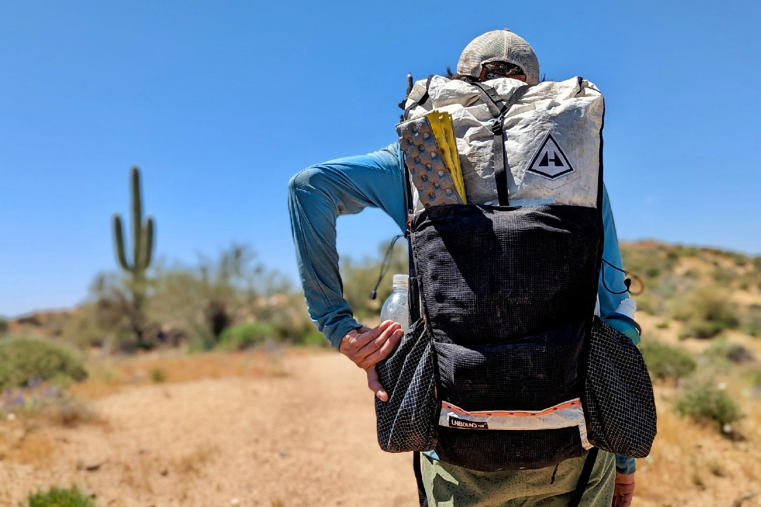 A hiker wearing the Hyperlite Mountain Gear Unbound 40 backpack on a desert trail - the hiker is reaching for a water bottle in the side pocket and theres a z seat in the front mesh pocket