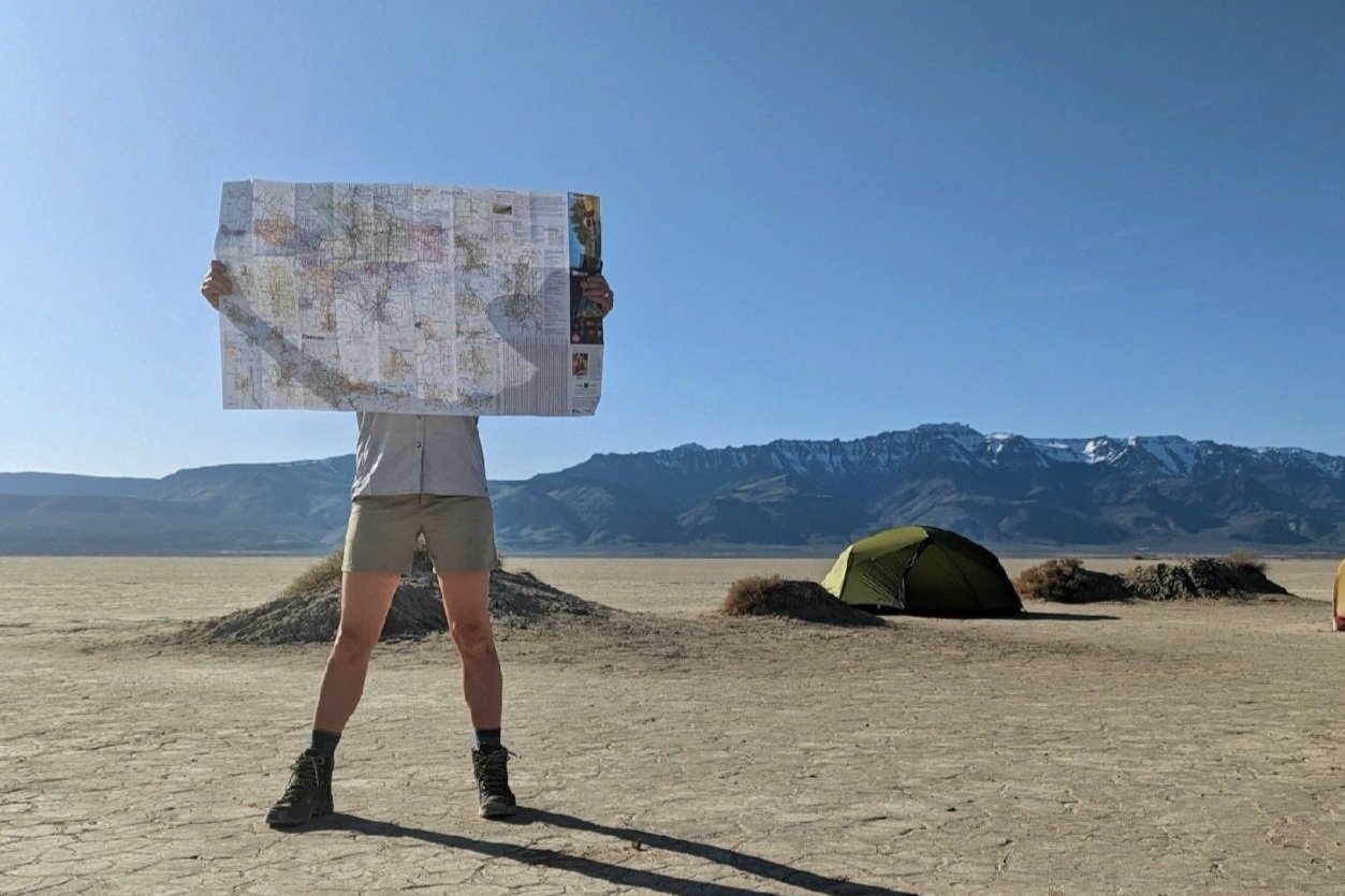 A hiker wearing the Patagonia Quandary Shorts holding up a map with some tents and mountains in the background