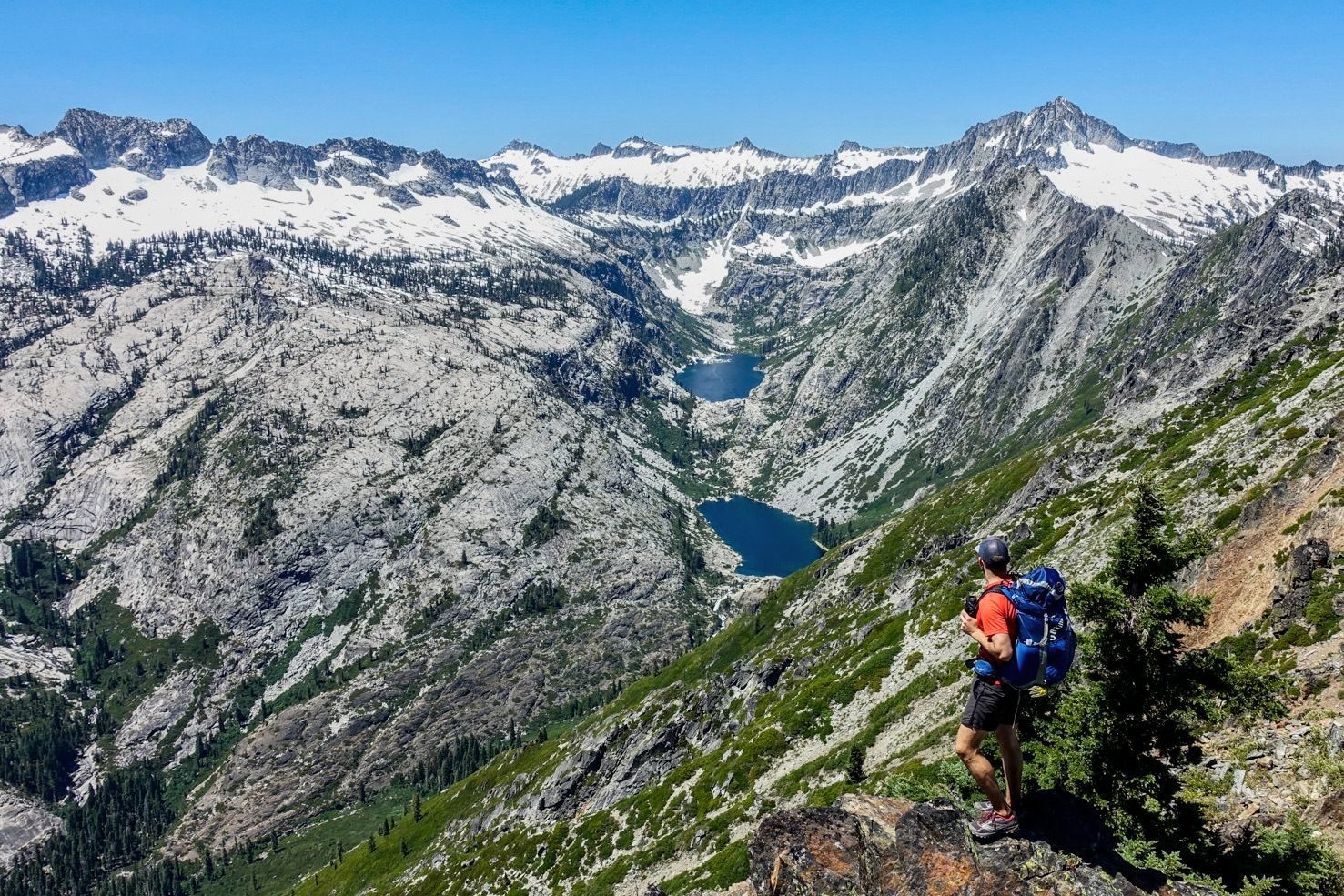 A backpacker wearing the Osprey Exos backpack on a trail overlooking a valley lined with snow-capped mountains