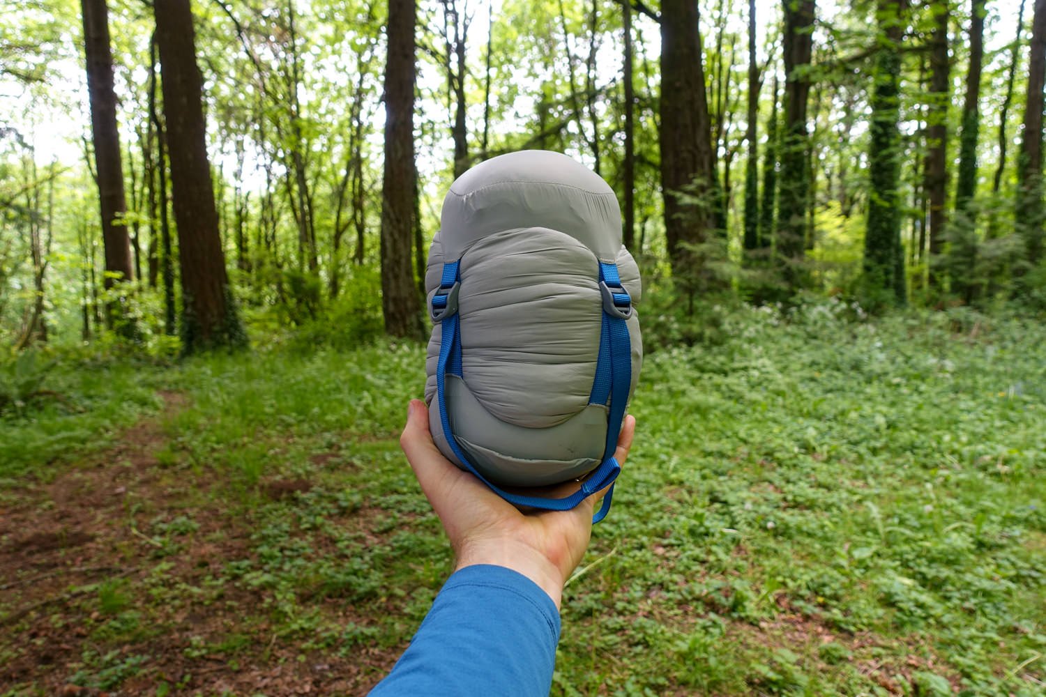 A hand holding up a backpacking sleeping bag in a compression sack in a wooded area