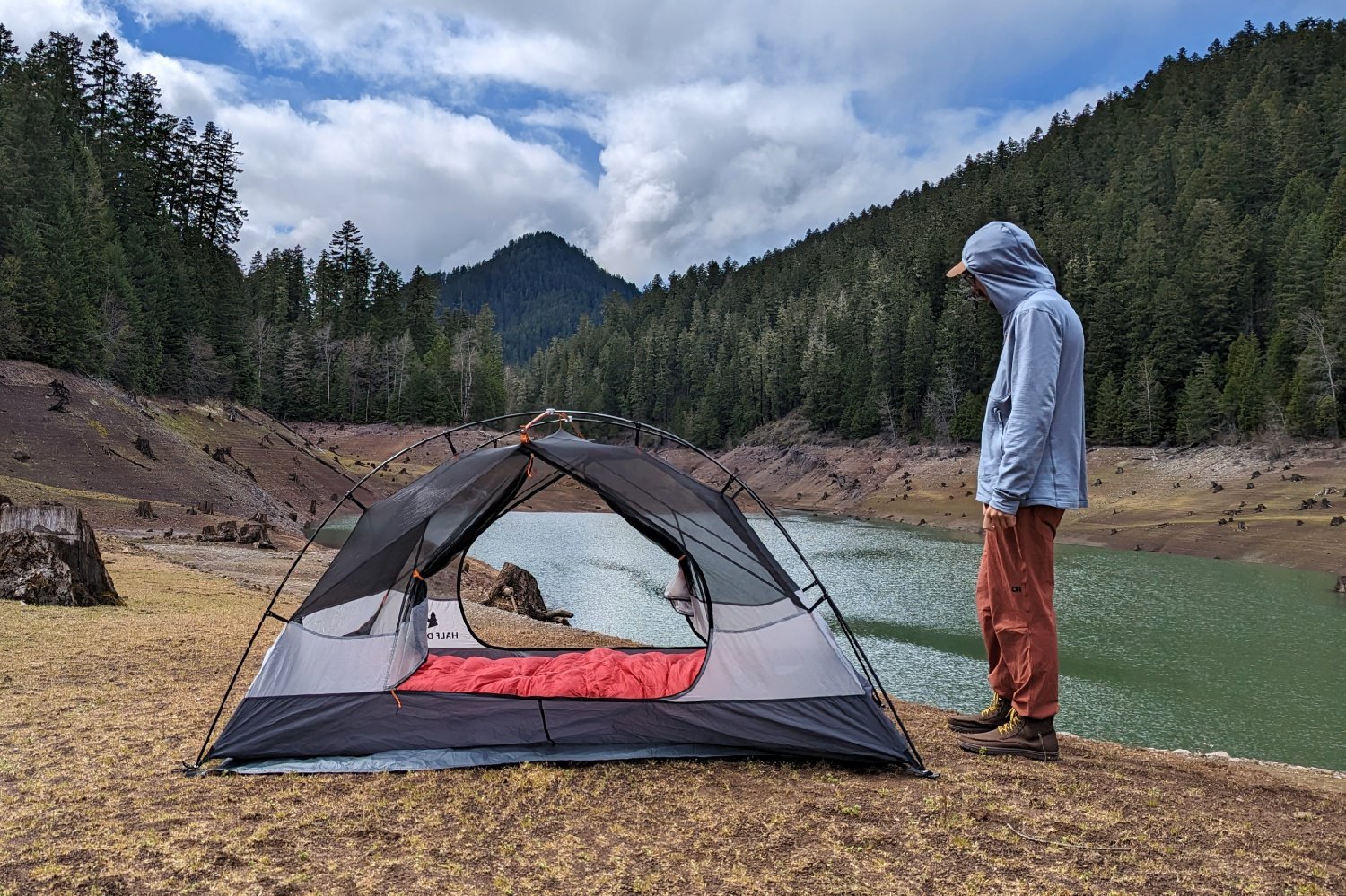 A hiker standing next to the set up  REI Half Dome 2 with the rainfly off and the doors open - you can see a red sleeping bag inside and there's a lake a mountain and a pine forest in the background