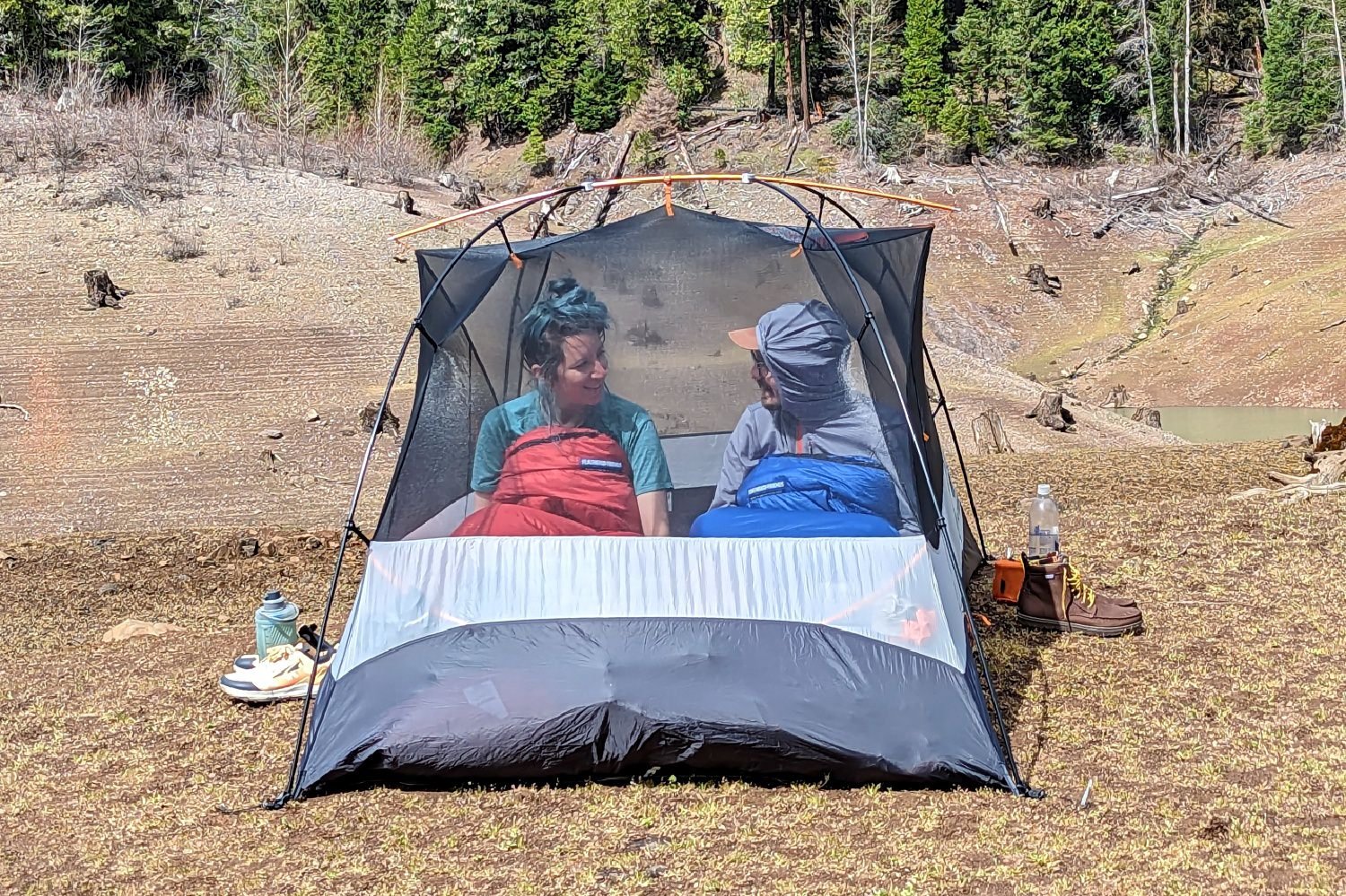 A view through the mesh body of the REI Half Dome 2 of two hikers sitting inside and smiling at each other with sleeping bags pulled up to their chests