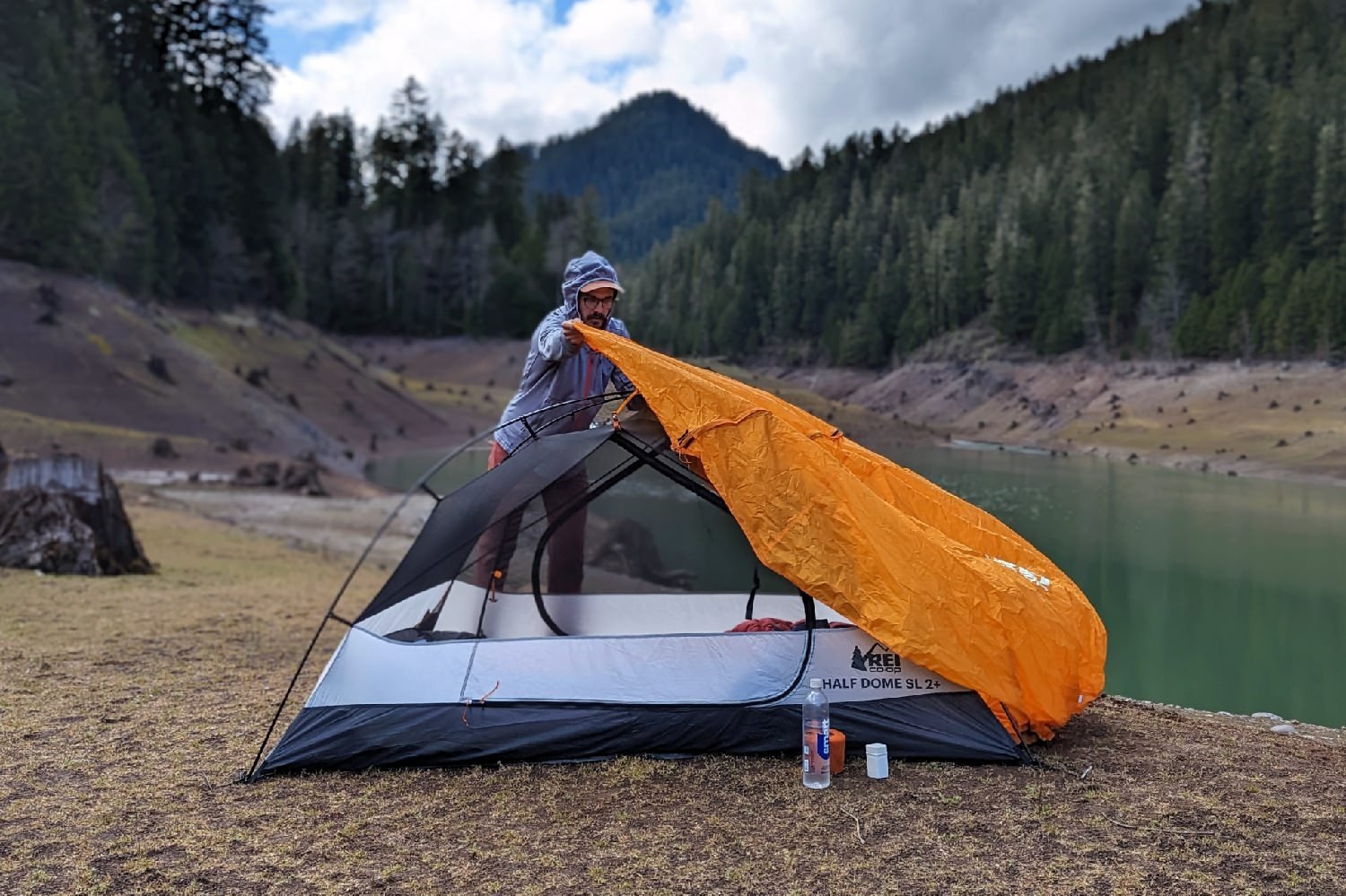 A hiker putting the rainfly on the REI Half Dome SL2 Tent with a lake and a mountain in the background