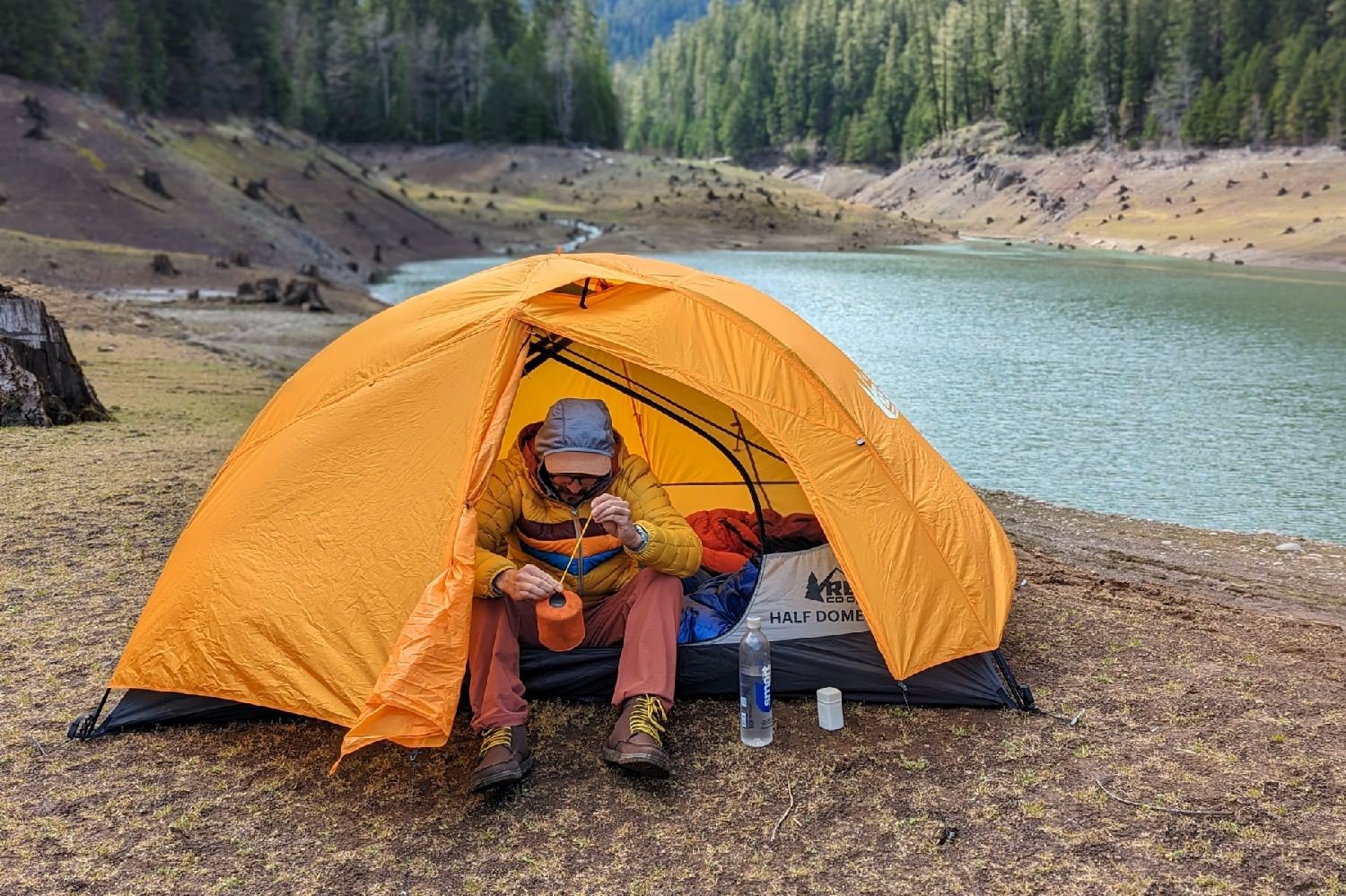 A hiker sitting inside the REI Half Dome 2 with the door and one side of the vestibule open - the hiker is putting away some gear and there's a lake and a pine forest in the background