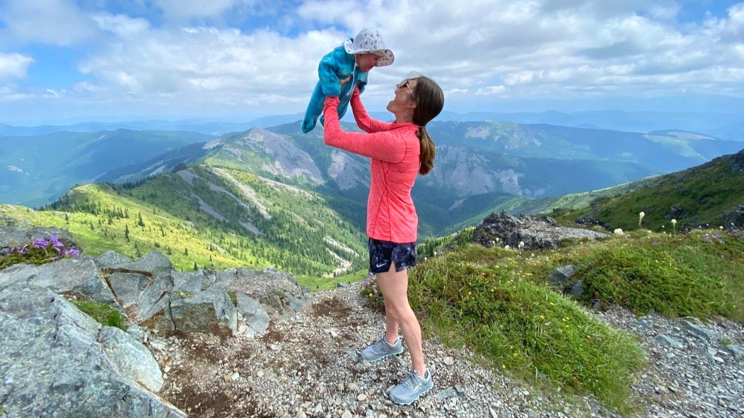 An outdoorsy mom holding her baby up on a mountain ridge