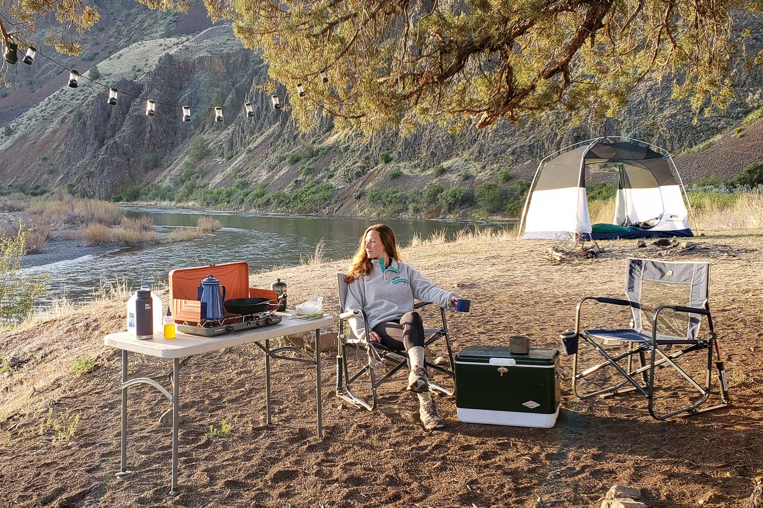 A woman sitting in the GCI Outdoor Freestyle Rocker Chair in a beautiful campsite near the river