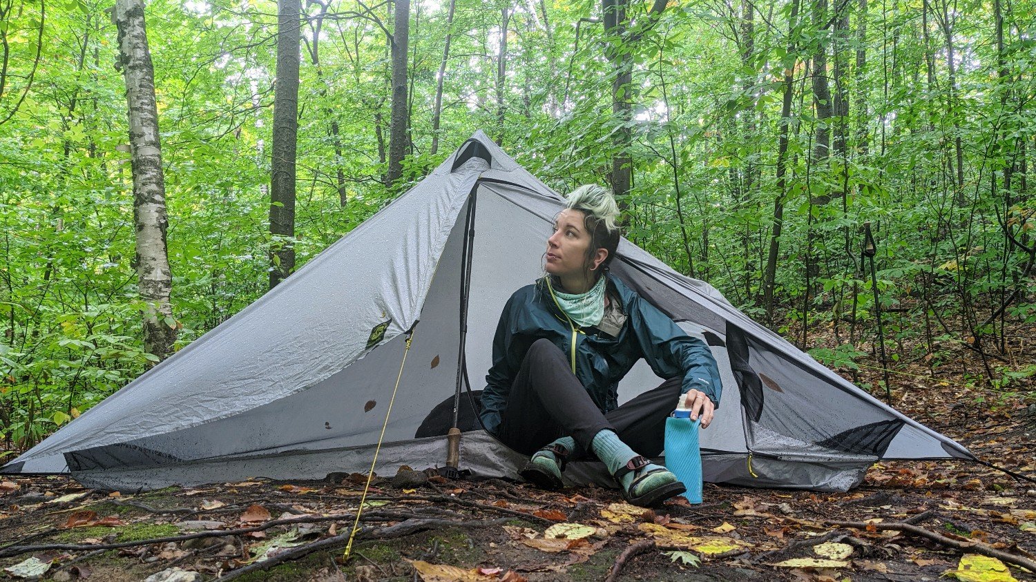 A hiker sitting in the doorway of the Six Moon Designs Lunar Solo at a campsite in a forest