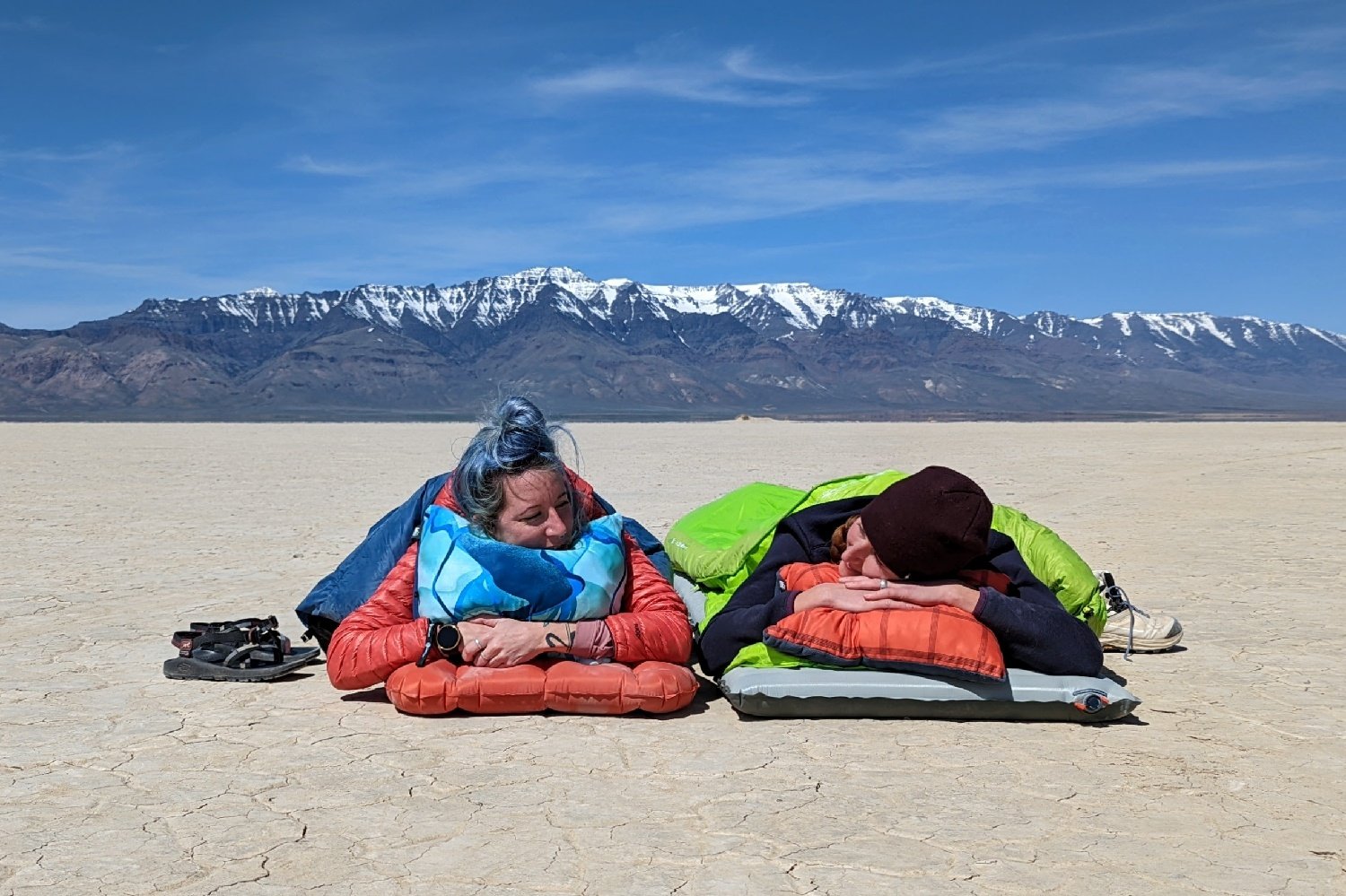 Two hikers laying on sleeping pads with backpacking quilts in the desert with mountains in the background