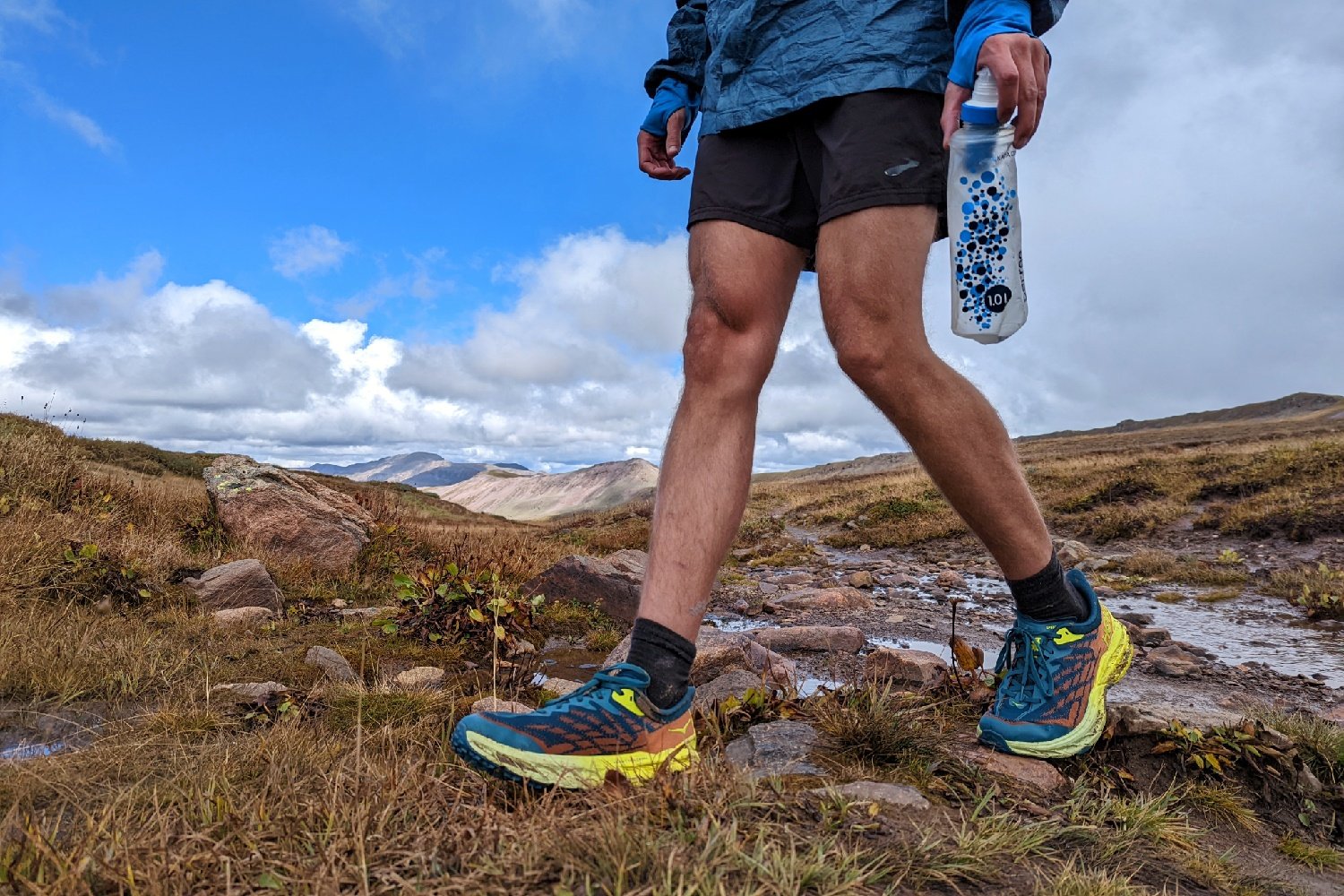 A hiker wearing Hoka Speedgoat hiking shoes on a trail with mountinas in the distance