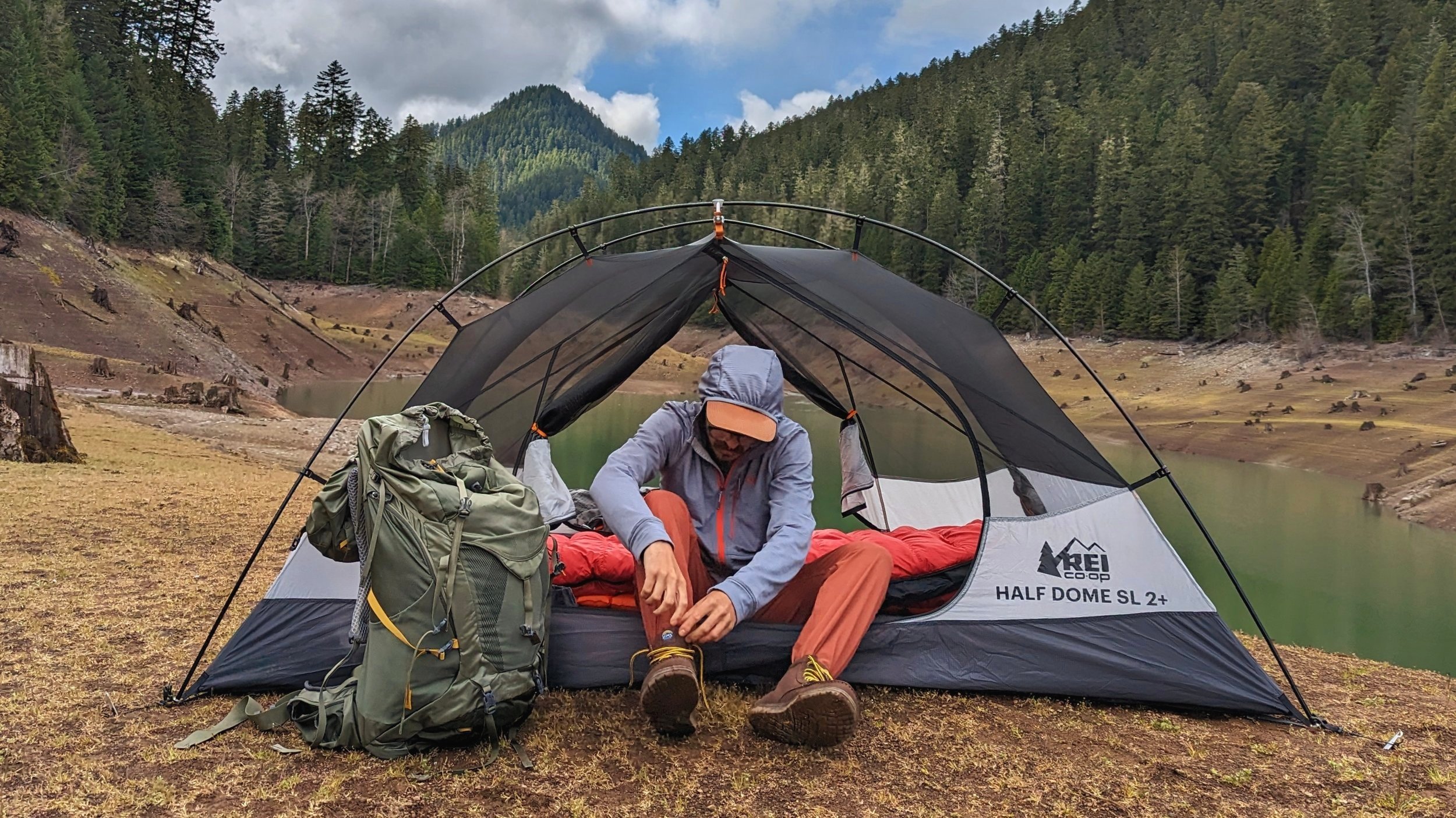 A hiker sitting in the open doorway of the REI Half Dome Sl 2 adjusting his shoes - there's a lake pine forest and a mountain in the background