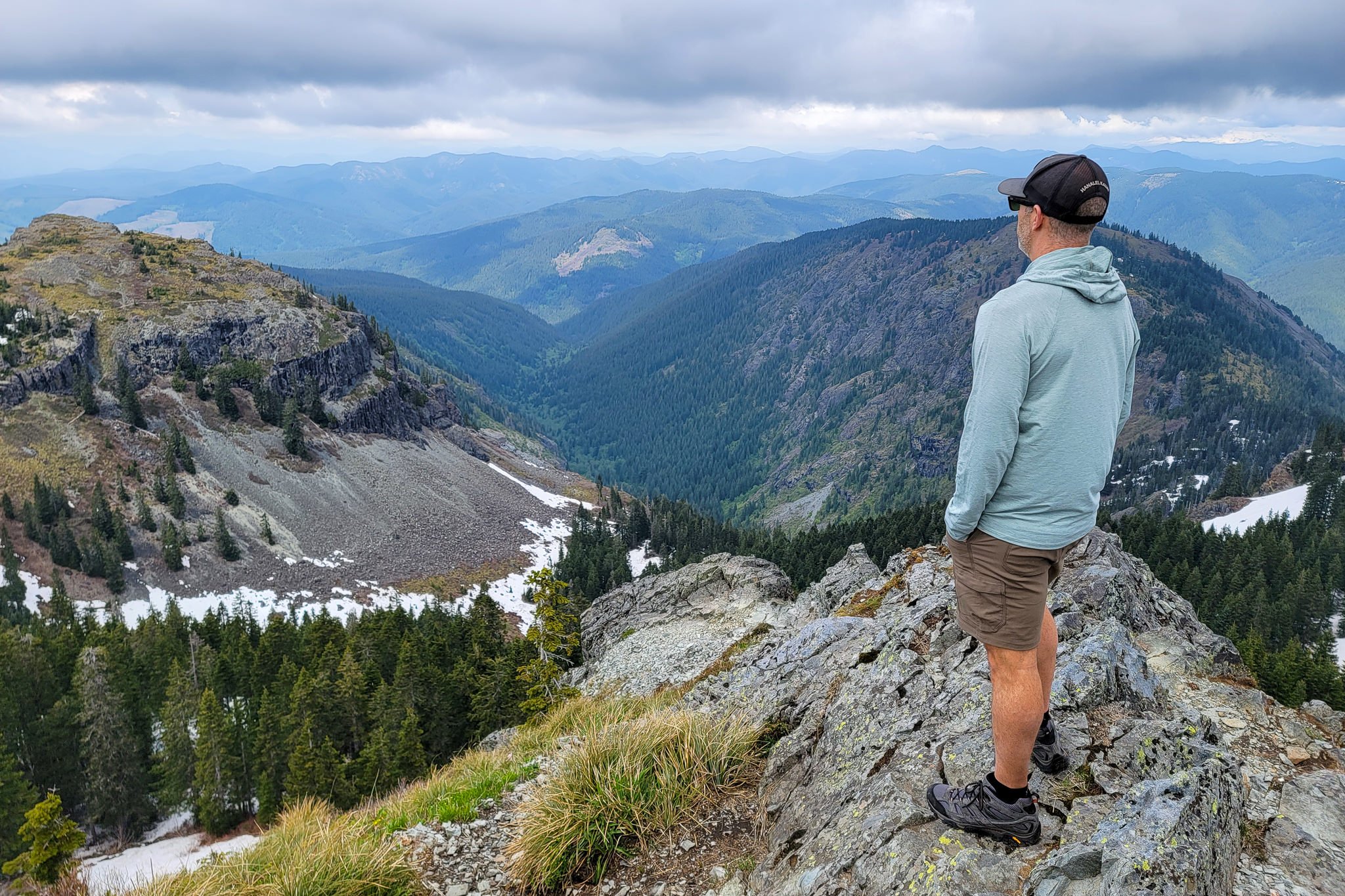 A hiker wearing Prana Stretch Zion II shorts standing at the top of a mountain with a view of a valley and other peaks