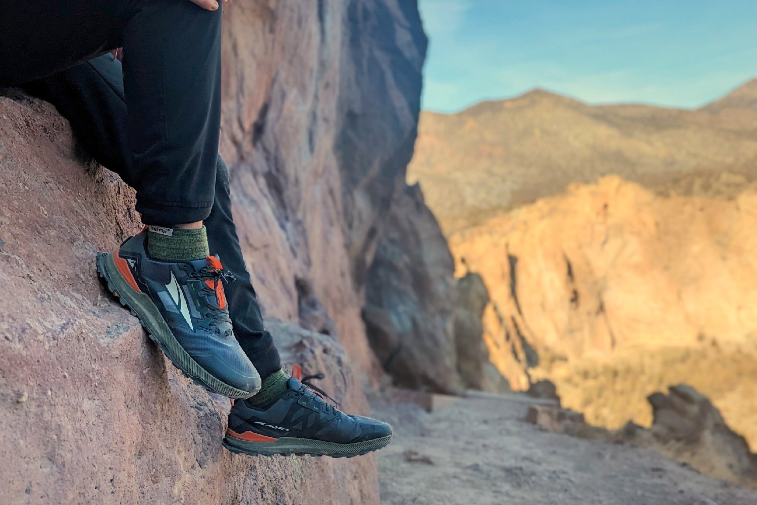 Hey a hiker wearing Altra Lone Peak hiking shoes, sitting on a rock with a trail and distant mountains in the background