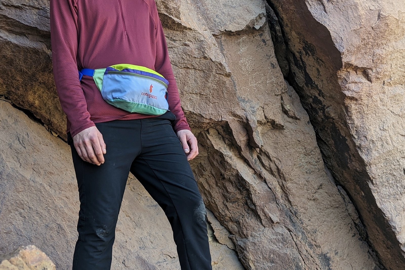 Waist level view of a hiker wearing the Cotopaxi Bataan fanny pack with a rocky wall in the background