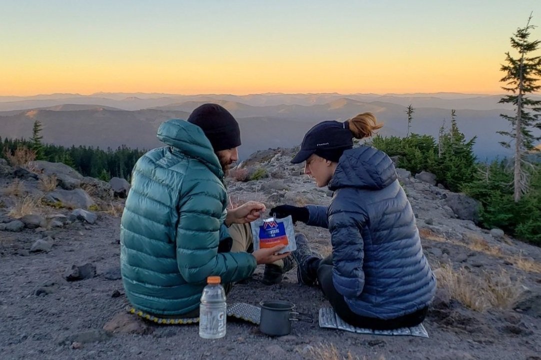 Two hikers sharing a freeze dried backpacking meal with a mountain view in the background