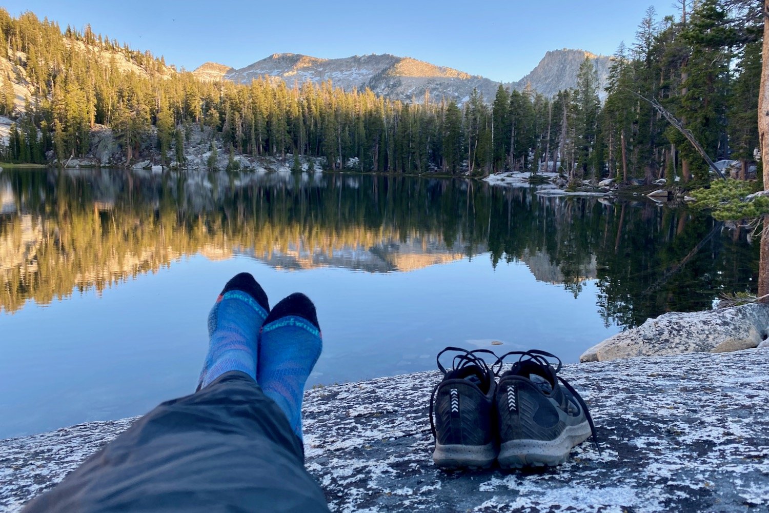 A hiker's feet with smartwool socks on in front of a lake mirroring some mountains