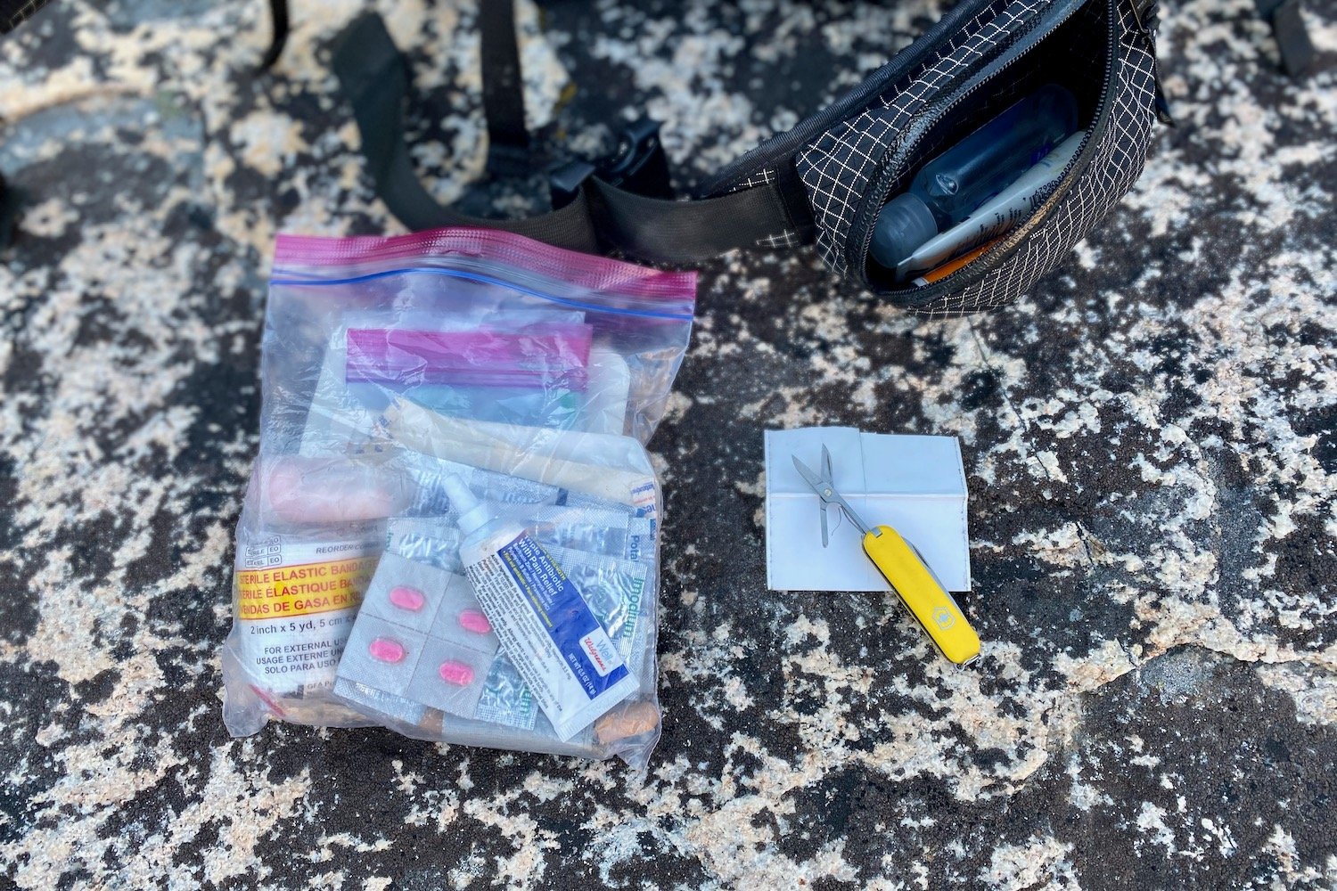 Top-down view of a DIY first aid kit in a Ziploc bag and a Swiss Army Classic multitool being used to cut tape